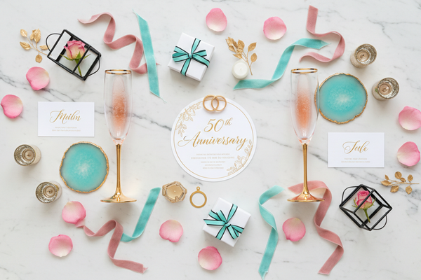 create an elegant flat lay for Wedding anniversary part on a light marble background. Shadowless with central focus point. Color palette: soft gold, white, lively rose pink, turquoise and black accents.