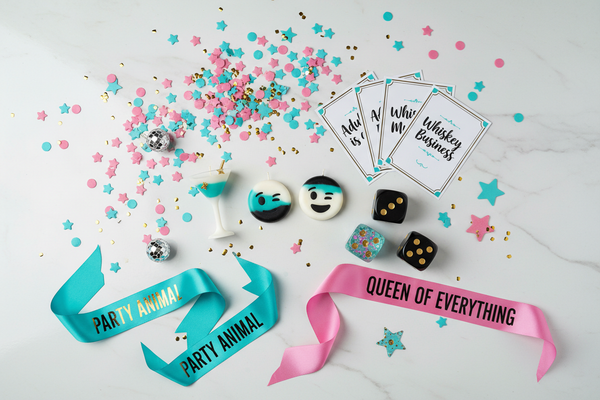create a stylish flat lay of playful adult party items for a "Grown-up fun" collection. Include humorous novelty party accessories such as cheeky shaped candles, playful confetti, joke cards, fun dice, sashes and party decorations. The mood should be fun, cheeky and light-hearted - never explicit vulgar. Color palette: turquoise, black, white, medium-saturated pink, gold accents. Place items on light cool off white background with subtle marble texture. Scattered confetti for energy. No birthday
