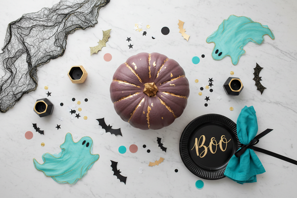 Flat lay stylish Halloween party decorations on a light marble background, one large statement pumpkin in the center surrounded by smaller bats, ghots, cobwebs, confetti and decorations, elegant black, dark dusty rose, turquoise and gold palette, playful but chic boutigue party styling, soft bright lightning, not too many tiny objects, no horror, no orange.