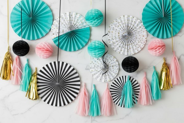 Playful yet elegant flat lay composition of hanging party decorations, including paper fans, honeycomb balls, tassels, spirals, visible vertical strings and ribbons, subtle vertical orientation suggesting hanging elements, soft white marble background, controlled festive mood, balanced playful arragement, high-end e-commerce photography. Color palette: turquoise, black, white, medium-saturated rose pink, gold accents.