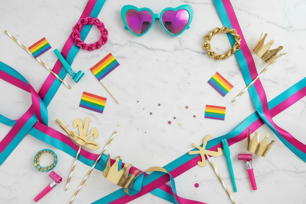 Flat lay Pride party decorations and accessories on a light marble background, mostly turquoise, pink and gold tones with only a few small rainbow flags, stylish festive decor, confetti, ribbons, accessories and party props, bold and lively mood, boutique party shop aesthetic, real objects, not pastel, not muted, no baby shower look, no unicorn style, no soft beige tones, no text, no clutter.