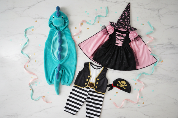 top view flat lay of three toddler party costumes. Cute and playful character costumes only. One dino, one fun witch and one pirate. Color palette mainly : turquoise, pink, white, black, gold. Light marble background with subtle ribbons and confetti. Clearly costumes, no pajamas, no normal outfits, no accessories. Clothing only, no models.