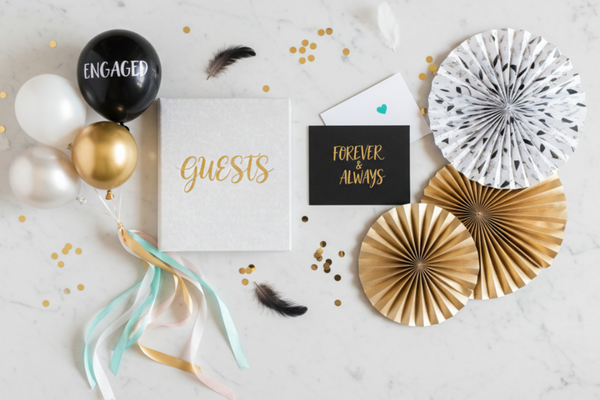 Create an elegant slightly asymmetrical flat lay for Wedding and angagement party on a light marble background. White and gold color palette with black, turquoise and pink accents. Shadowless with center focus point. Add guest book, greeting card, balloons, ribbons, paper fans.